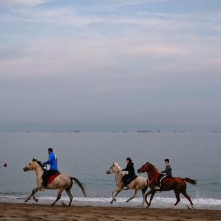 Ryttare galopperar längs en strand medan oljetankrar och lastfartyg står i kö vid Hormuzsundet. 11 mars 2026. 