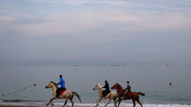 Ryttare galopperar längs en strand medan oljetankrar och lastfartyg står i kö vid Hormuzsundet. 11 mars 2026. 